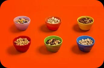 Cashews, pistaccios, and mixed nuts are displayed in colorful bowls against an orange surface.